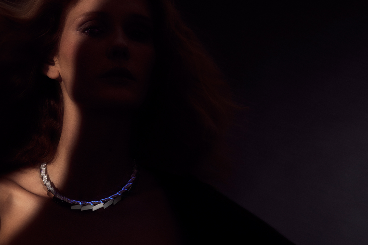 This is a close up dramatic color photo of a woman model wearing the Light Halo Necklace. She is mostly obscured in shadow with the platinum necklace glowing blue in black light. The design is by artist Sardwell for Renisis.