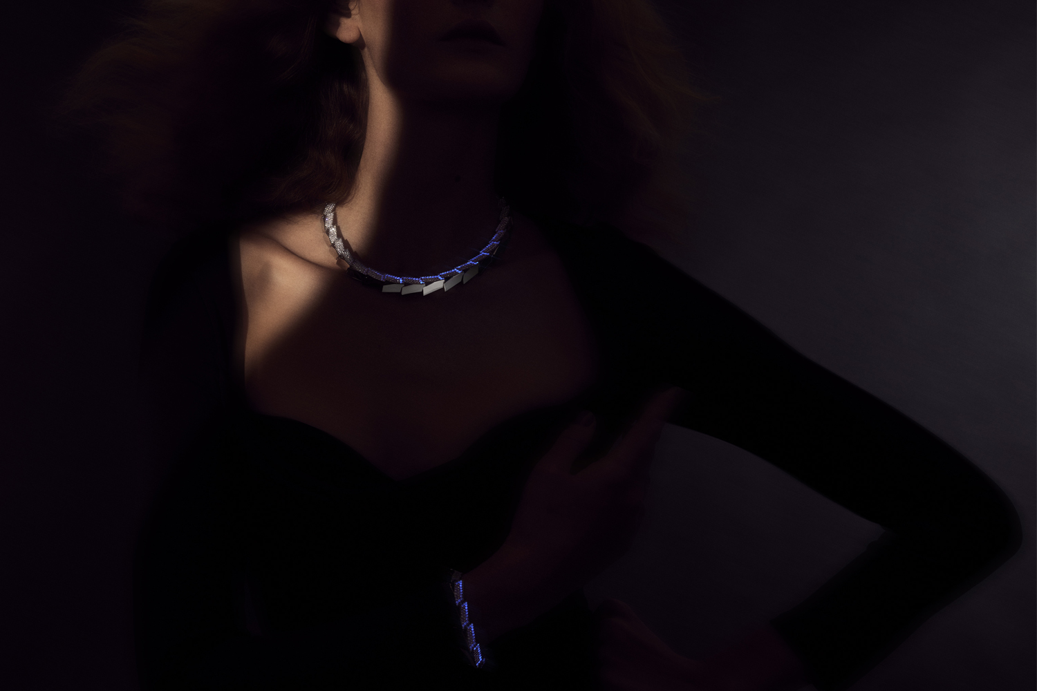 This is a dramatic color photo of a woman model wearing the Light Halo Necklace and bracelet. She is mostly obscured in shadow with the platinum necklace and bracelet glowing blue in black light.
