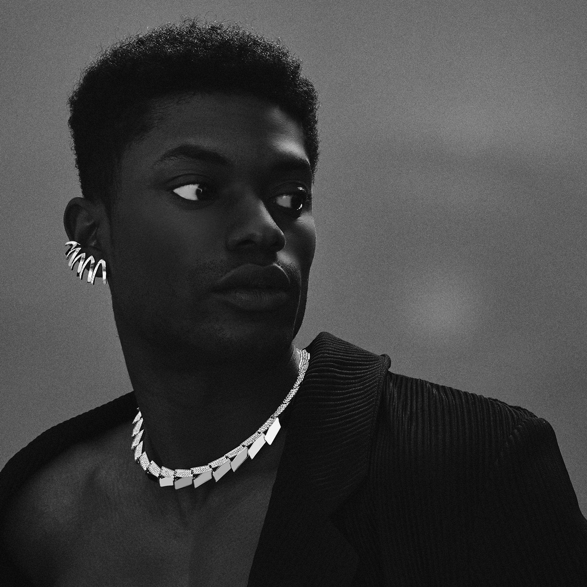 This is a black and white photo of a male model wearing two pieces of Renisis jewelry, the Light Halo necklace and the Echo Chamber Ear Cuff designed by Sardwell.